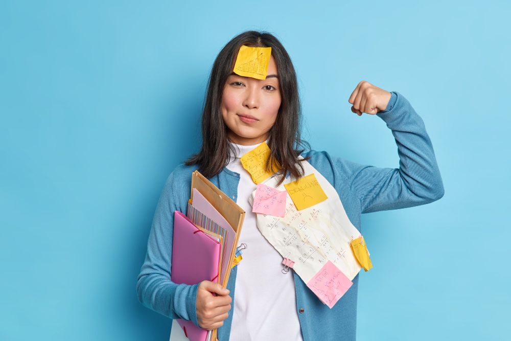 powerful female student raises arm shows muscles feels confident after working diploma paper wears stickers forehead holds folders